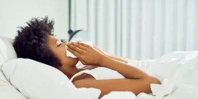 Una mujer con el cabello rizado tumbada en una cama sonándose la nariz con un pañuelo de papel. 
