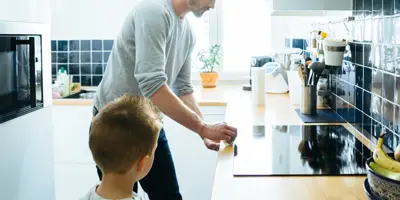 Un hombre con un suéter gris limpiando la cocina junto a su hijo pequeño.