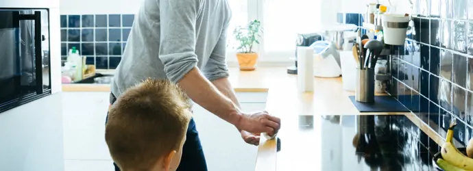 Un hombre con un suéter gris limpiando la cocina junto a su hijo pequeño.