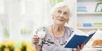 Una señora con el pelo blanco sonriendo con una taza de té y un libro.