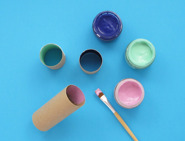 Três rolos de papel higiénico apoiados em bancada azul ao pé de copos de tinta coloridos e pincel.