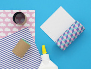 Dois rolos de papel higiénico apoiados em bancada azul com papel de embrulhos em cores diferentes e tubo de cola.