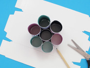 Tubos de papel higiénico decorados apoiados em base de caixa de cartão com lápis e tesoura ao lado.