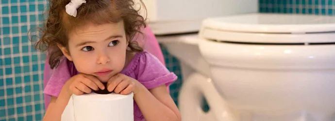 Una niña sentada en un baño mirando pensativa con un rollo de papel higiénico.