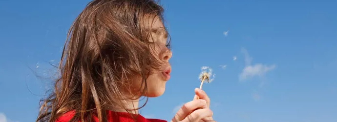Una niña que podría necesitar algunos remedios caseros para la fiebre del heno mientras sopla un diente de león con un cielo azul de fondo