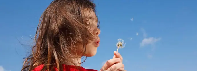 Uma criança pequena que pode precisar de alguns remédios caseiros para a febre dos fenos enquanto sopra um dente-de-leão com um céu azul de fundo