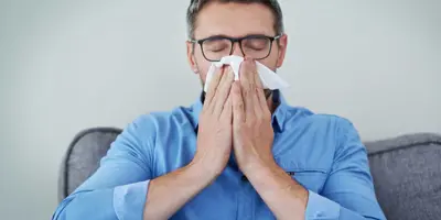 Um homem de óculos e camisa azul sentado num sofá a assoar o nariz