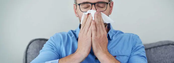Um homem de óculos e camisa azul sentado num sofá a assoar o nariz