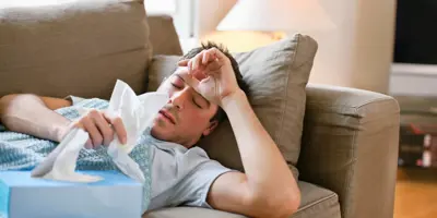 Homem deitado no sofá com gripe enquanto segura lenços de papel