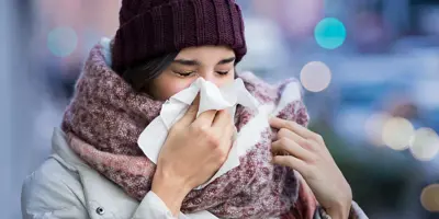 Mulher com roupas de inverno volumosas a sofrer de sintomas de sinusite e a espirrar num lenço de papel ao ar livre