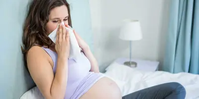 Uma mulher grávida sentada numa cama a assoar o nariz com um lenço de papel