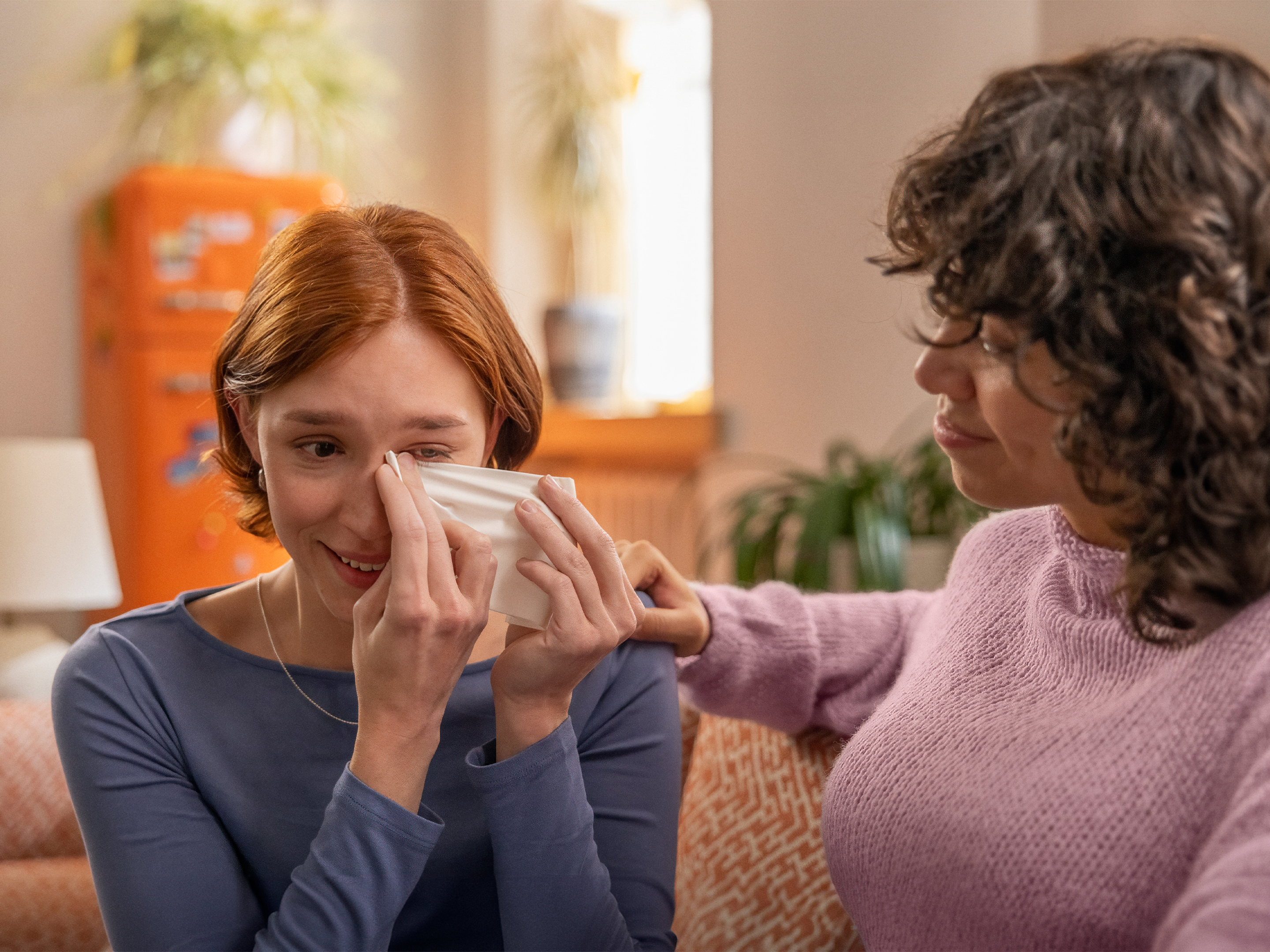 Una mujer se limpia los ojos con un pañuelo mientras otra a su lado la consuela en una sala luminosa y acogedora.