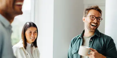 Três pessoas, uma delas com uma caneca de café na mão, sorriem numa sala iluminada.