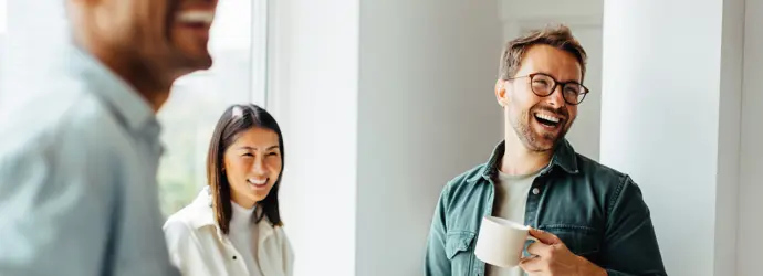 Três pessoas, uma delas com uma caneca de café na mão, sorriem numa sala iluminada.